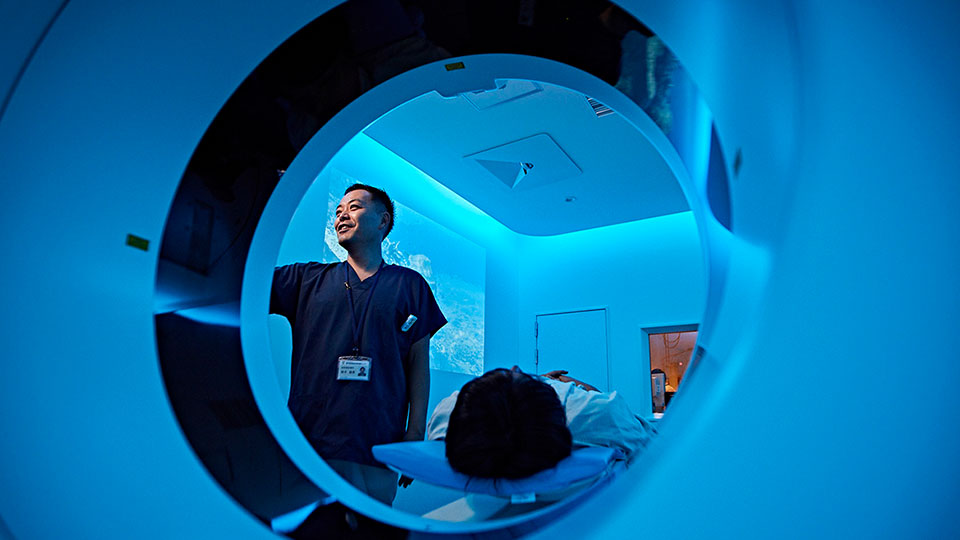 Photo of clinician setting up a scan at the CT scanner&rsquo;s gantry, with the patient on the CT system&rsquo;s patient table 